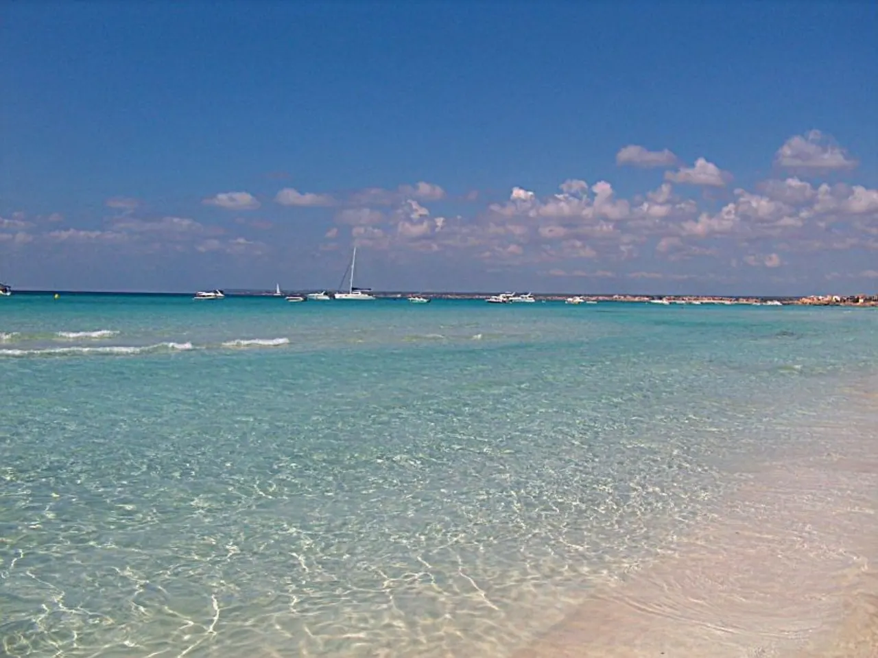 The clear, pale-turquoise shallows of Es Trenc over white sand, boats on the horizon