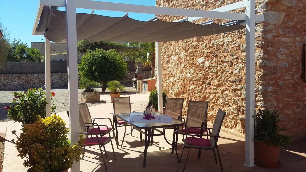 An outdoor dining table and chairs under the white pergola against the dry-stone wall, by day